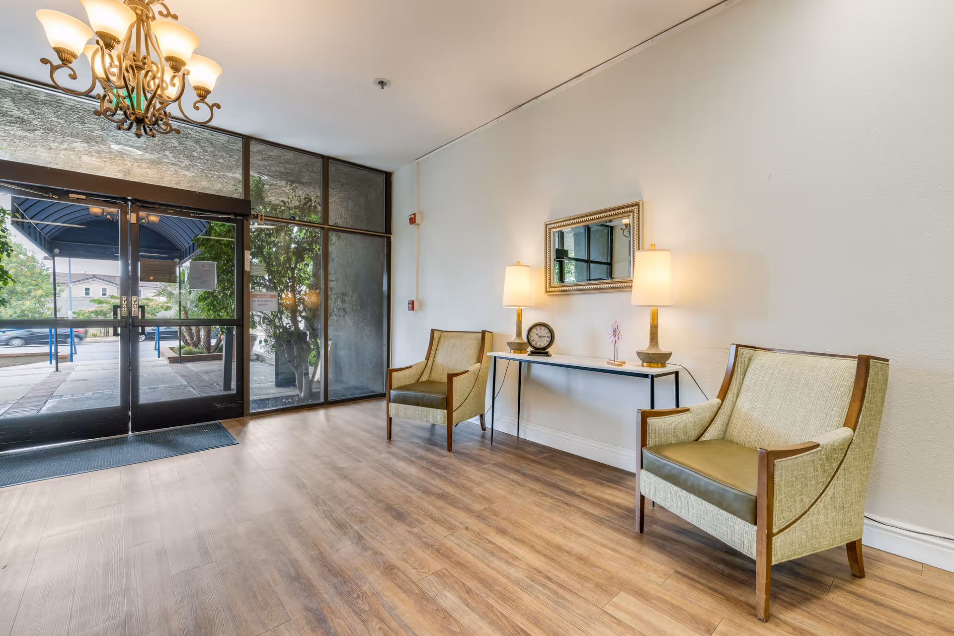 Entrance area of a facility with glass double doors leading outside. Inside, there are two beige armchairs with wooden frames placed on either side of a narrow console table. The table holds two lamps, a small clock, and a decorative item. A rectangular mirror hangs on the wall above the table. The floor is wooden, and a chandelier with multiple lights hangs from the ceiling.