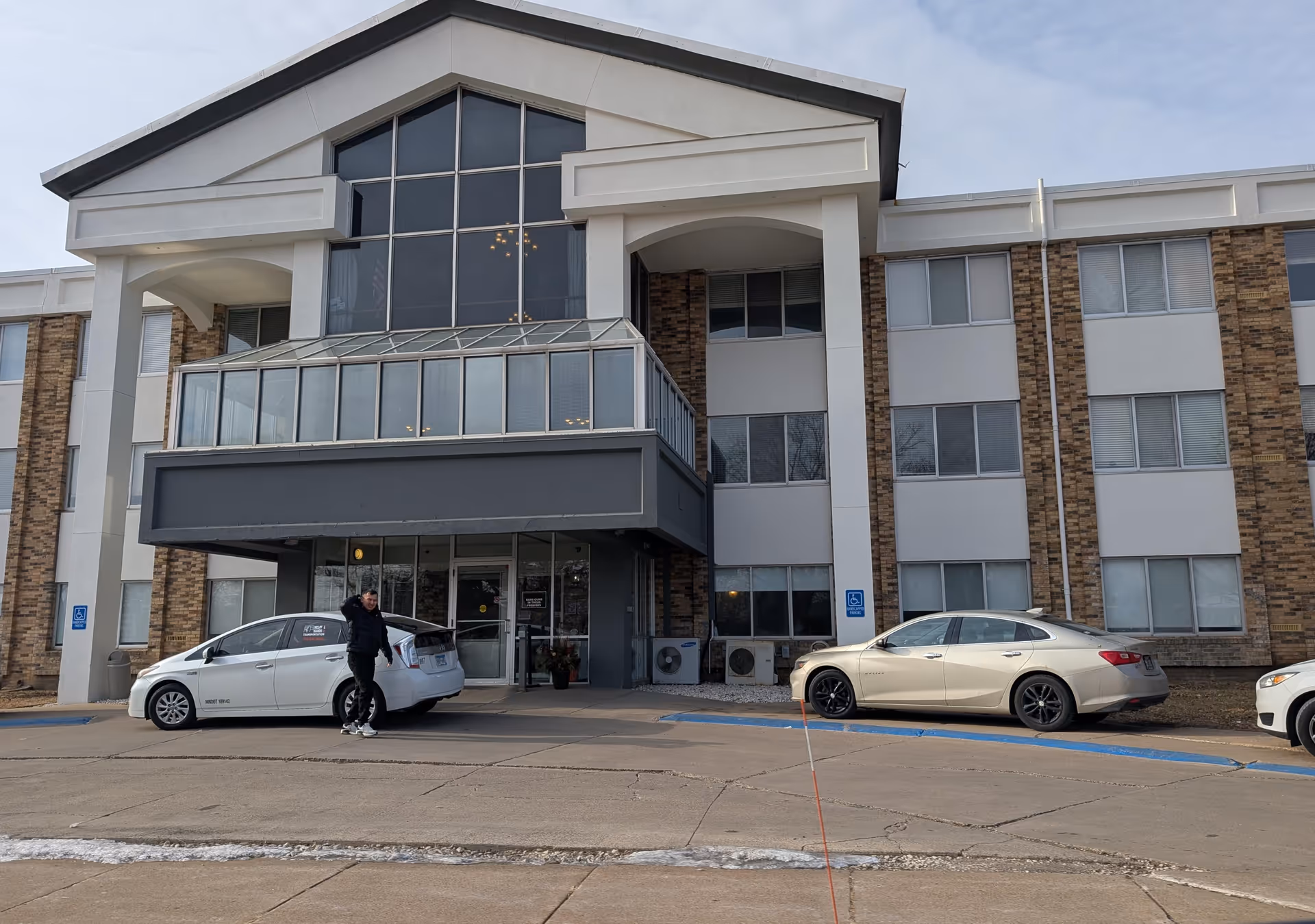 Front exterior view of a multi-story senior living facility named Edenbrook Edina with large windows and a covered entrance. Two cars are parked in front, one white and one beige, and a person is walking near the white car. The building has a combination of brick and white paneling.
