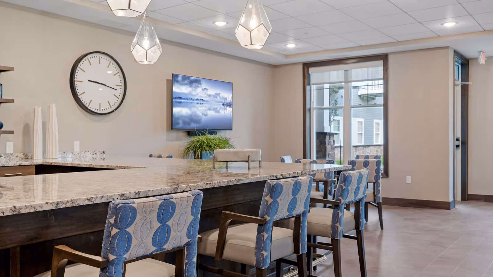 Communal dining area with a long marble-topped island, patterned upholstered chairs, pendant lights, and a wall-mounted TV.