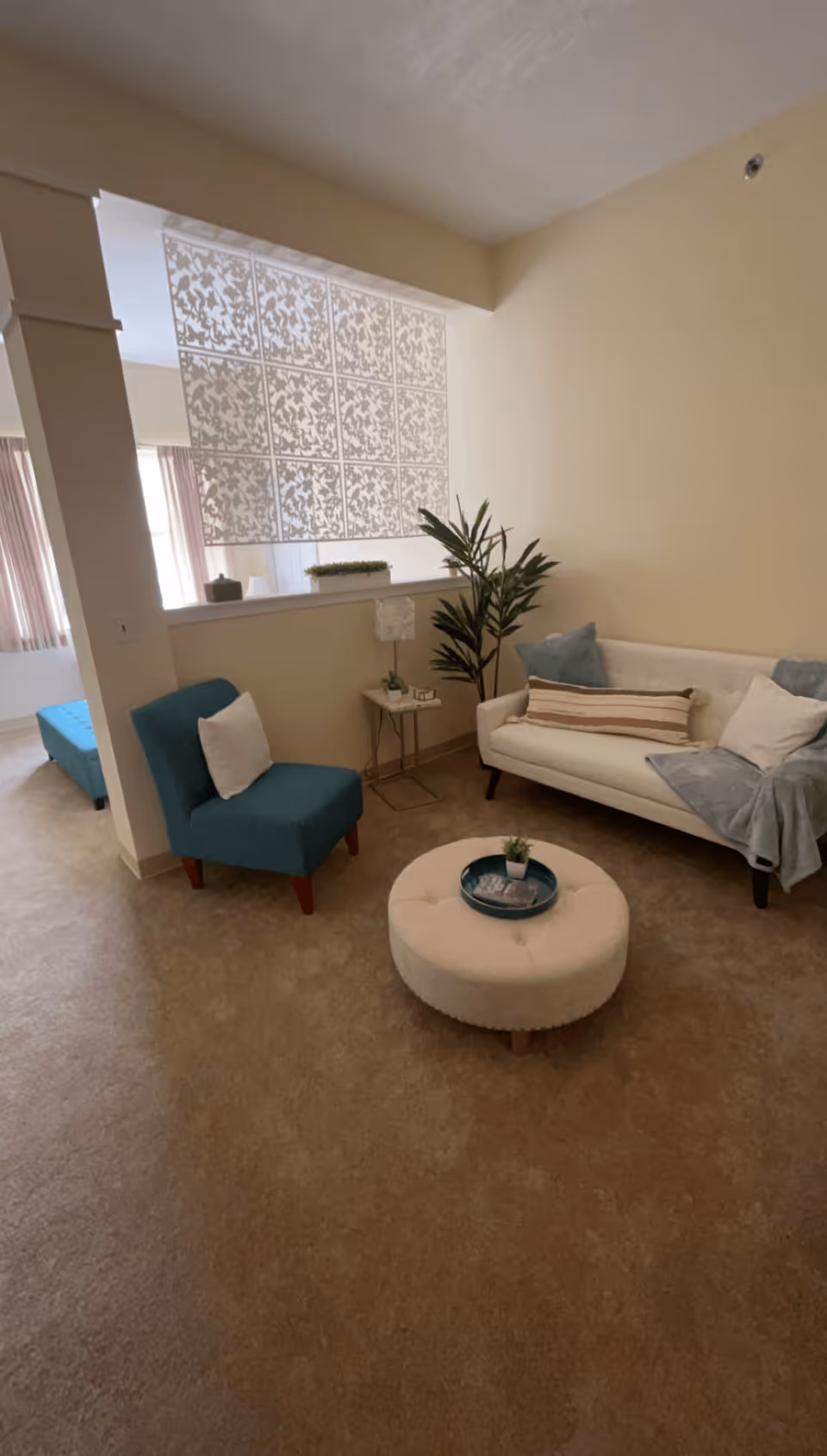 A cozy living room area with a white sofa adorned with pillows and a throw blanket, a teal armchair with a white pillow, a round white ottoman with a small plant and tray on top, a small side table with a lamp and decorative items, and a tall green plant in the corner. The room has beige walls and carpet, and a decorative glass block partition partially separates the space from another area with windows and curtains.