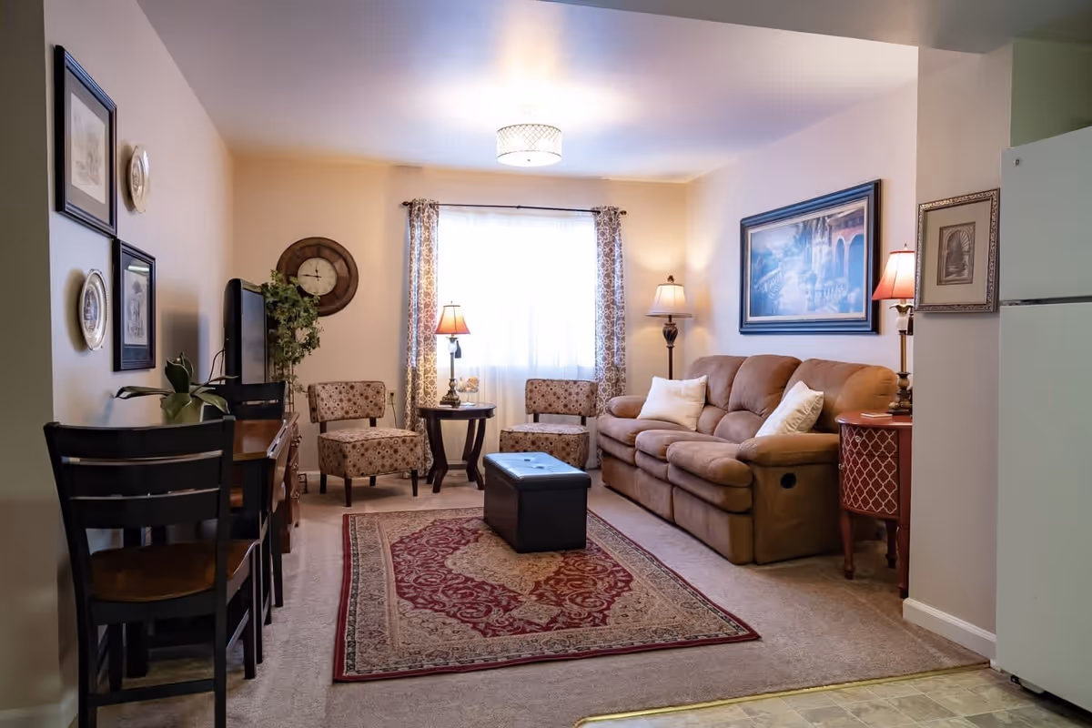 A cozy living room with a brown sofa adorned with white pillows, two patterned armchairs, a dark wooden coffee table with storage, and a red and beige patterned rug. The room is softly lit by two table lamps and a floor lamp, with a large window covered by sheer curtains and patterned drapes. There is a TV on a wooden stand, a wall clock, framed artwork, and a small dining area with two chairs and a table. A white refrigerator is partially visible on the right side.