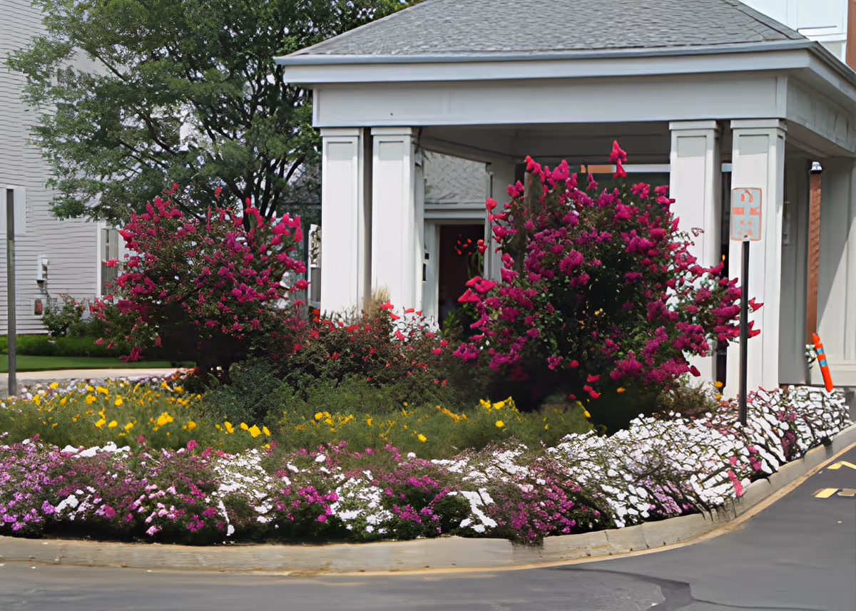 Flower beds and shrubs in front of a covered entrance with white columns to a building.