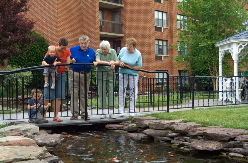 Two elderly women, one elderly man, a woman, and two young children standing on a small black metal bridge over a pond with fish, in front of a brick residential building with balconies and surrounded by greenery.