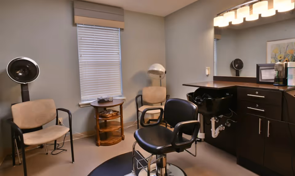 Interior of a small salon room with a black salon chair in front of a black sink and dark wood cabinetry. Two beige chairs and a wooden side table are placed near a window with closed blinds. The room has light gray walls and a large mirror with lights above the sink area.