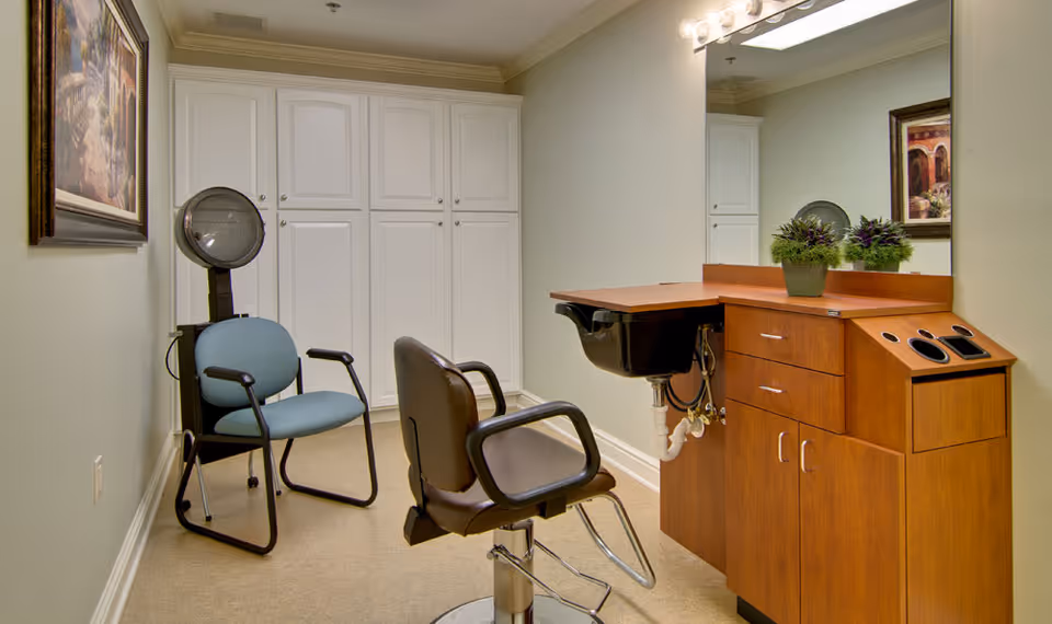 Interior view of a hair salon area in a senior living facility with a salon chair, a blue waiting chair, a hair dryer, a wooden cabinet with a sink, a large mirror, and framed artwork on the walls.