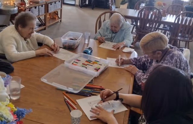 Four elderly women sitting around a wooden table engaged in coloring and drawing activities with colored pencils and markers in a well-lit room with wooden chairs and shelves in the background.