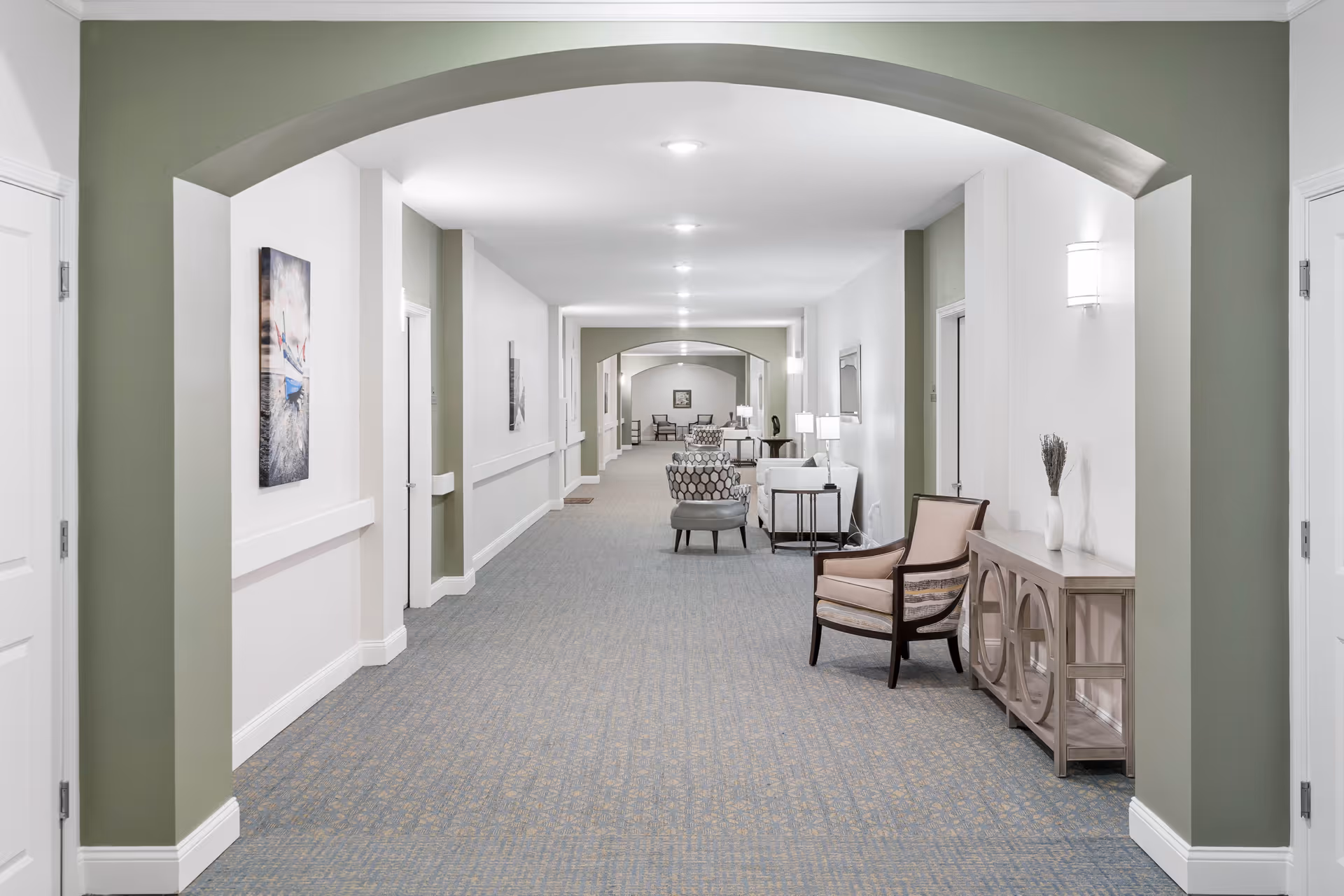 A long, well-lit hallway in a senior living facility with light green and white walls, carpeted floor, and multiple seating areas featuring chairs and small tables with lamps. Decorative wall art and a console table with a vase are also visible.