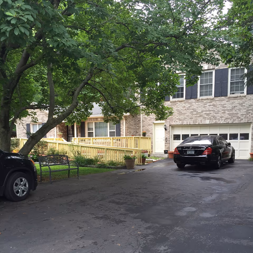 Exterior view of a residential building with a brick facade, black shutters, and a double garage door. There is a black car parked in the driveway and a black SUV partially visible on the left. A large tree with green leaves provides shade over the driveway, and a wooden ramp with railings leads to the front door. A metal bench and potted plants are visible near the ramp.