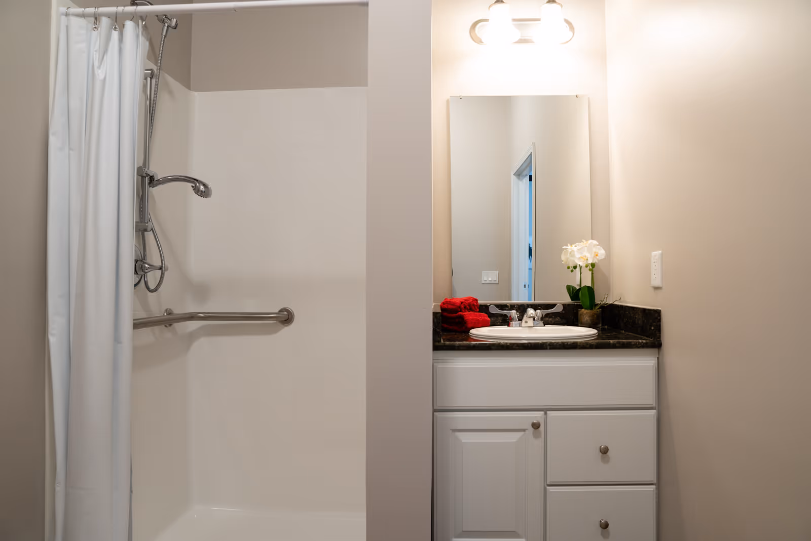 A clean bathroom with a white shower area featuring a handheld showerhead and a safety grab bar. Next to the shower is a white vanity with a dark countertop, a sink, a mirror, and a light fixture above. Red towels and a small white orchid plant are placed on the countertop.