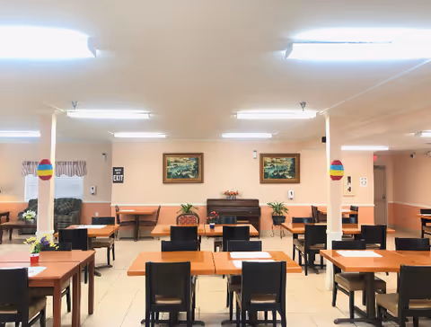 Dining room with multiple square tables and chairs arranged in rows, a piano and framed paintings on the back wall.