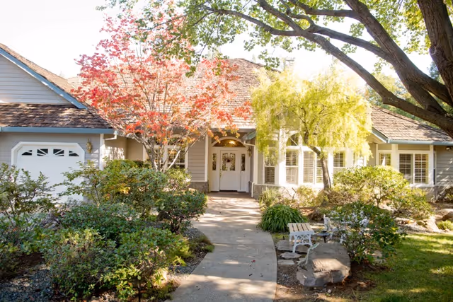 Front entrance of a single-story house with a paved walkway, landscaped shrubs and trees, a bench, and an attached garage.