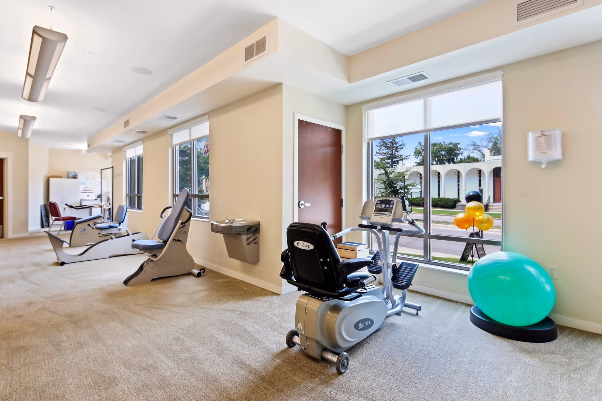 A bright exercise room with large windows showing an outdoor view. The room contains several pieces of exercise equipment including stationary bikes and a recumbent bike, as well as a large green exercise ball on a stand. The walls are light-colored and there is a water fountain mounted on one wall.