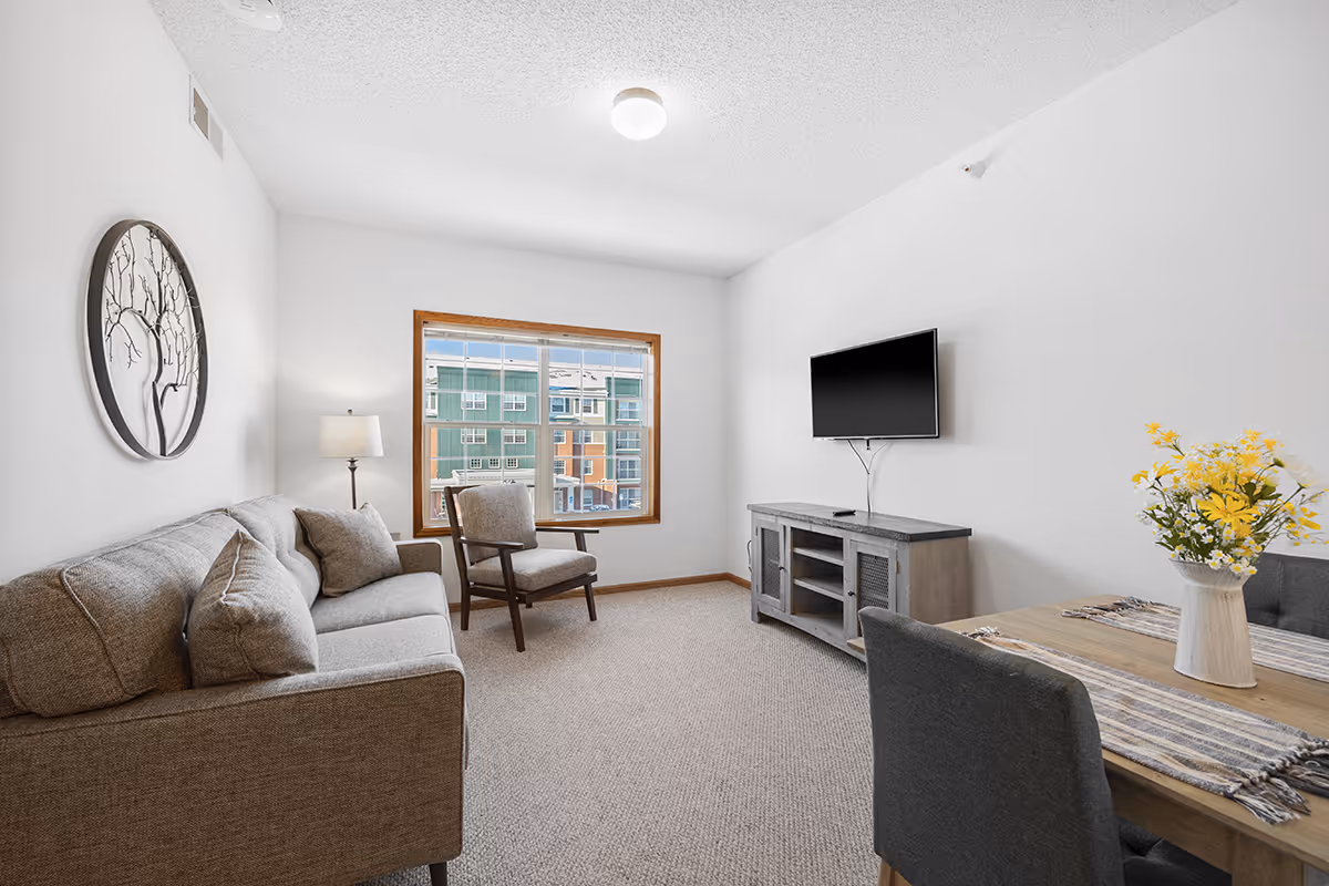 Bright living room with a sofa, armchair, wall-mounted TV, window overlooking neighboring buildings, and a small dining table with a vase of flowers.