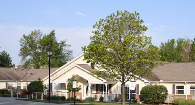 Single-story beige senior living building front with a covered porch, lawn, trees, and a small parking area with handicap signs.