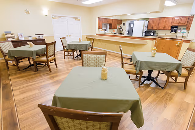 A dining area in an assisted living facility with several square tables covered with green tablecloths, each table surrounded by wooden chairs with patterned cushions. The room has wood flooring, beige walls, and a kitchen area with wooden cabinets and appliances visible in the background.