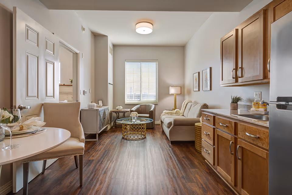A cozy senior living apartment interior featuring a small dining area with a round table and upholstered chairs on the left, a living area with a beige sofa, armchair, glass coffee table, and a floor lamp near a window with blinds. On the right side, there is a kitchenette with wooden cabinets, a countertop, and a refrigerator. The room has wood flooring and neutral-colored walls with framed artwork.
