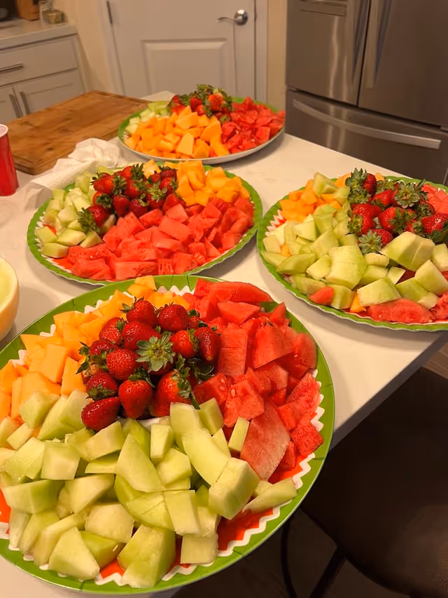 Four plates on a kitchen counter filled with an assortment of fresh fruit including strawberries, watermelon, cantaloupe, and honeydew melon. In the background, there is a refrigerator and a closed door.