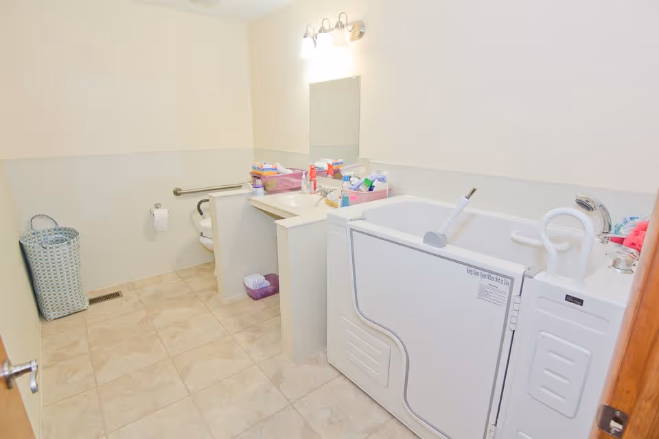 A clean, accessible bathroom with beige tiled floor and light-colored walls. The room features a walk-in bathtub with safety handles, a toilet with a grab bar, a vanity with a sink, mirror, and overhead light, and a laundry basket in the corner. Various toiletries and supplies are organized on the vanity counter.