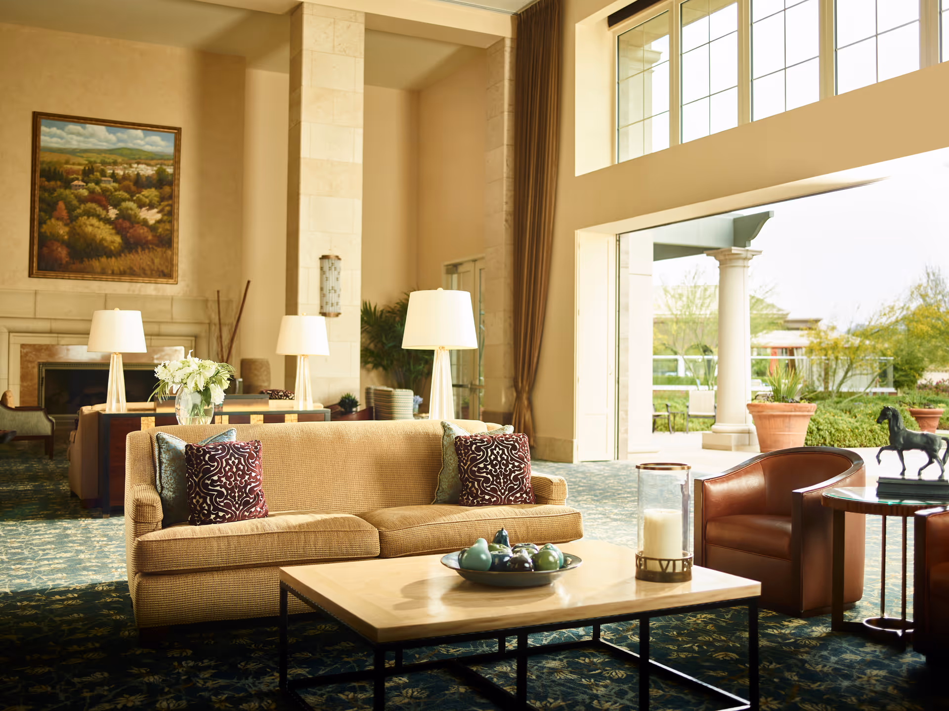 A spacious and well-lit living room area with a beige sofa adorned with decorative pillows, a wooden coffee table with a decorative bowl and candle, two brown leather armchairs, and large windows letting in natural light. The room features a fireplace, floor lamps, a large painting on the wall, and an open view to an outdoor garden area with potted plants and columns.