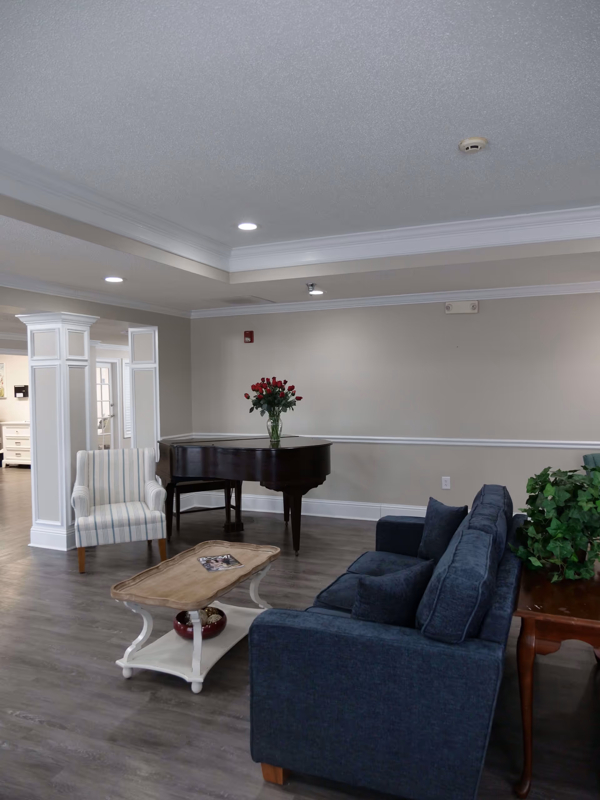A cozy living room area with a dark blue sofa, a wooden coffee table with a magazine on it, a striped armchair, and a black grand piano with a vase of red roses on top. The room has light-colored walls, recessed ceiling lights, and wood flooring.