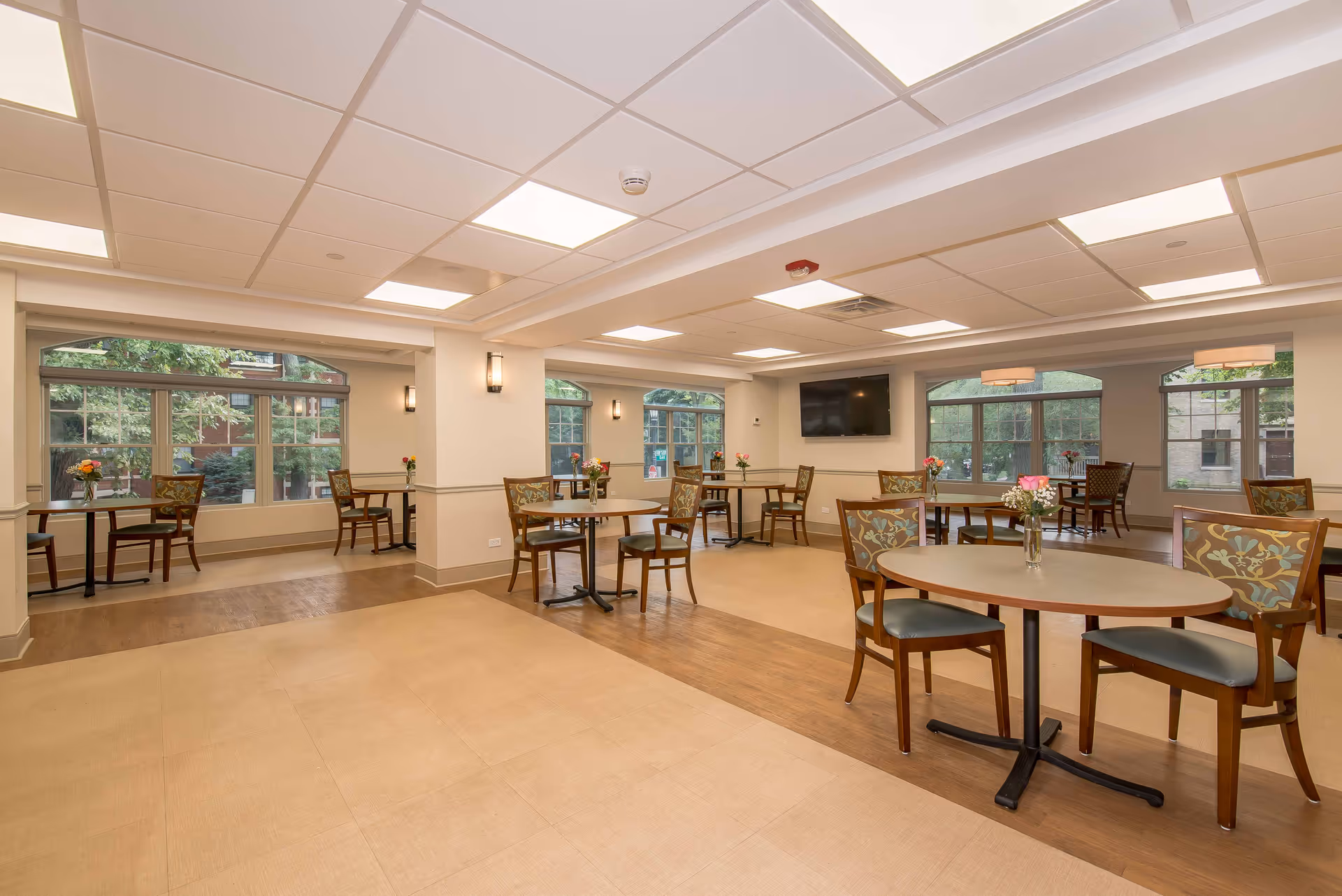 A spacious dining room with multiple round tables and chairs arranged neatly. Each table has a small vase with flowers. Large windows allow natural light to fill the room, and a flat-screen TV is mounted on one wall. The ceiling has recessed lighting panels, and the floor features a combination of light and dark wood tones.