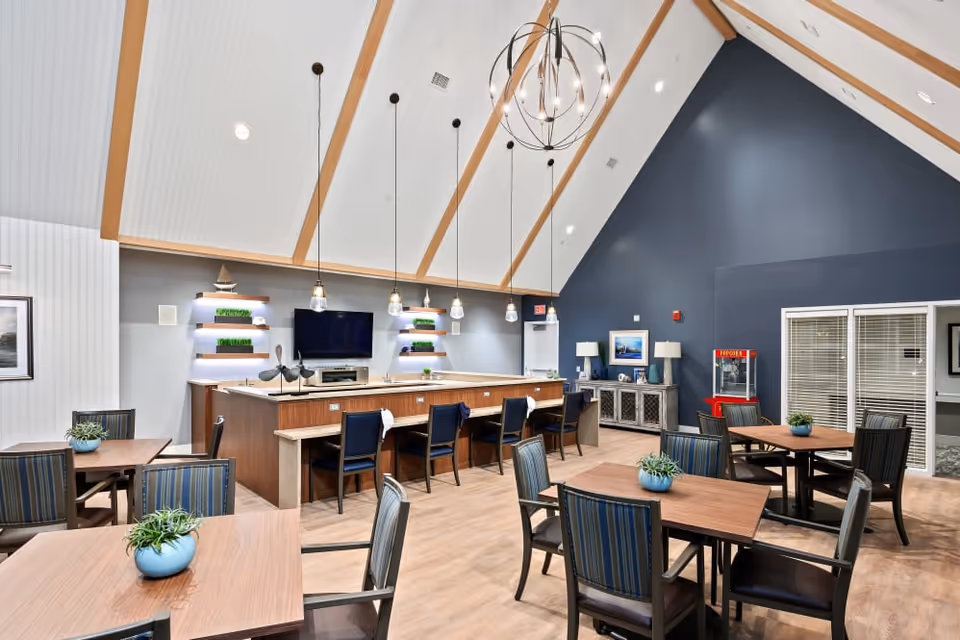 A spacious dining area with several wooden tables and striped cushioned chairs. The room features a high vaulted ceiling with wooden beams and modern pendant lights hanging above a central counter with bar stools. A large flat-screen TV is mounted on the wall behind the counter, flanked by shelves with decorative plants. The walls are painted in shades of blue and white, and there is a popcorn machine in the corner near a sideboard with lamps and framed pictures.