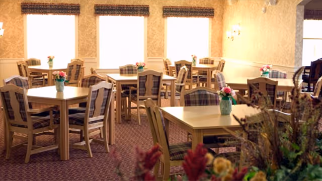 A dining room with several wooden tables and chairs arranged neatly. Each table has a small vase with pink flowers. The room has patterned wallpaper, carpeted flooring, and three large windows letting in natural light. There are wall sconces providing additional lighting.