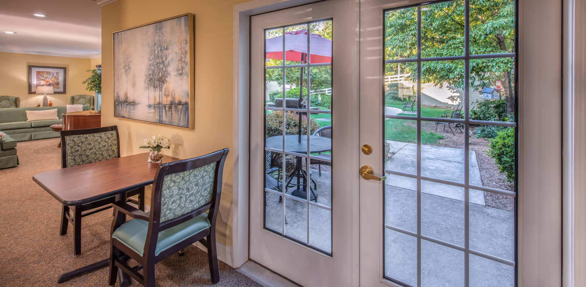 Interior view of a senior living facility showing a small wooden table with two chairs next to a glass door leading to an outdoor patio area with a table and umbrella. In the background, there is a living room area with green sofas, a side table with a lamp, and framed artwork on the walls.