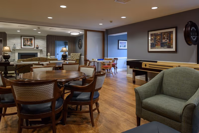 A cozy common area in a senior living facility featuring a round wooden table with six chairs, a green upholstered armchair, a shuffleboard table, and a fireplace with lamps and framed artwork on the walls. The room has wooden flooring and recessed ceiling lights.