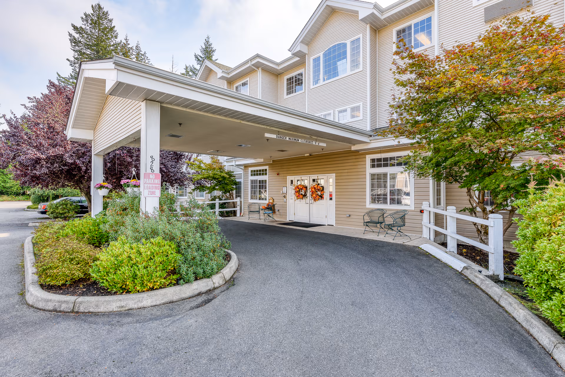 Covered driveway and entrance leading to the front doors of a multi-story assisted living building with landscaping and wreaths on the doors.