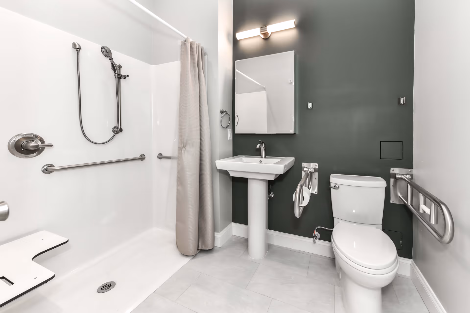 A modern, accessible bathroom featuring a walk-in shower with a handheld showerhead and grab bars, a white pedestal sink with a mirror and light above it, and a toilet equipped with safety rails on both sides. The walls are painted in a combination of dark green and white, and the floor is tiled in light gray.