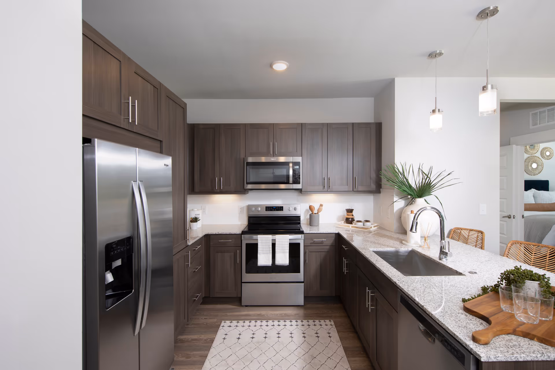 Modern kitchen with stainless steel refrigerator, oven, microwave, and dishwasher. The kitchen features dark wood cabinets, a light granite countertop with a sink, pendant lights hanging from the ceiling, and a small rug on the wooden floor. There are decorative plants and kitchen utensils on the counter, and a glimpse of a bedroom is visible through an open doorway.