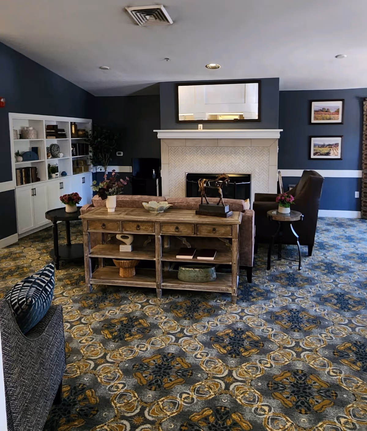A cozy senior living room with patterned carpet, a wooden console table with decorative items, a fireplace with a large mirror above it, a bookshelf filled with books and decor, and comfortable seating including a sofa and armchairs. The walls are painted dark blue with white trim, and framed pictures hang on the wall.