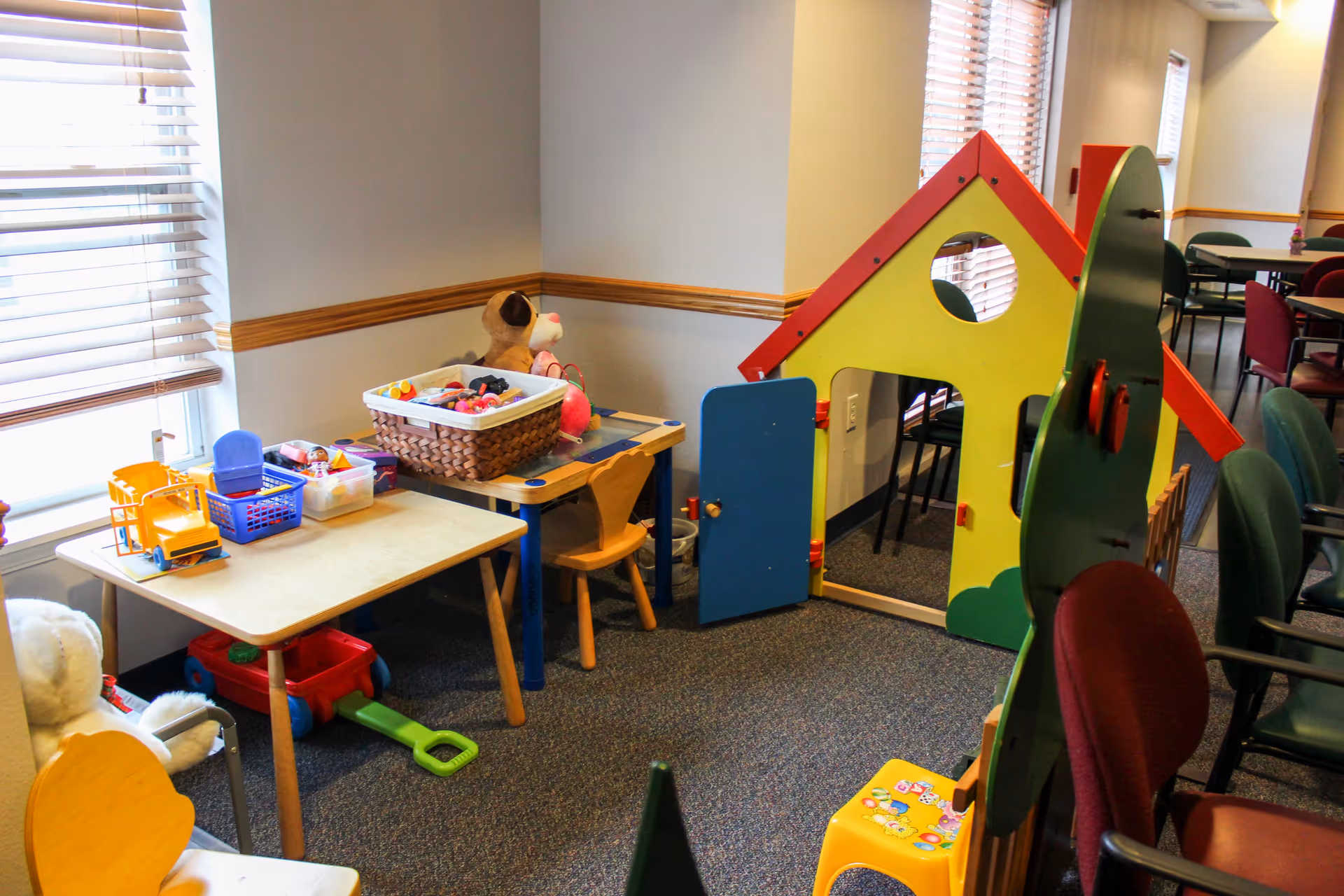 A small play area inside a senior living facility with children's toys, a small table with chairs, a basket of toys, a large teddy bear, and a colorful playhouse with a blue door. The area is next to a window with blinds and adjacent to a dining area with tables and chairs.
