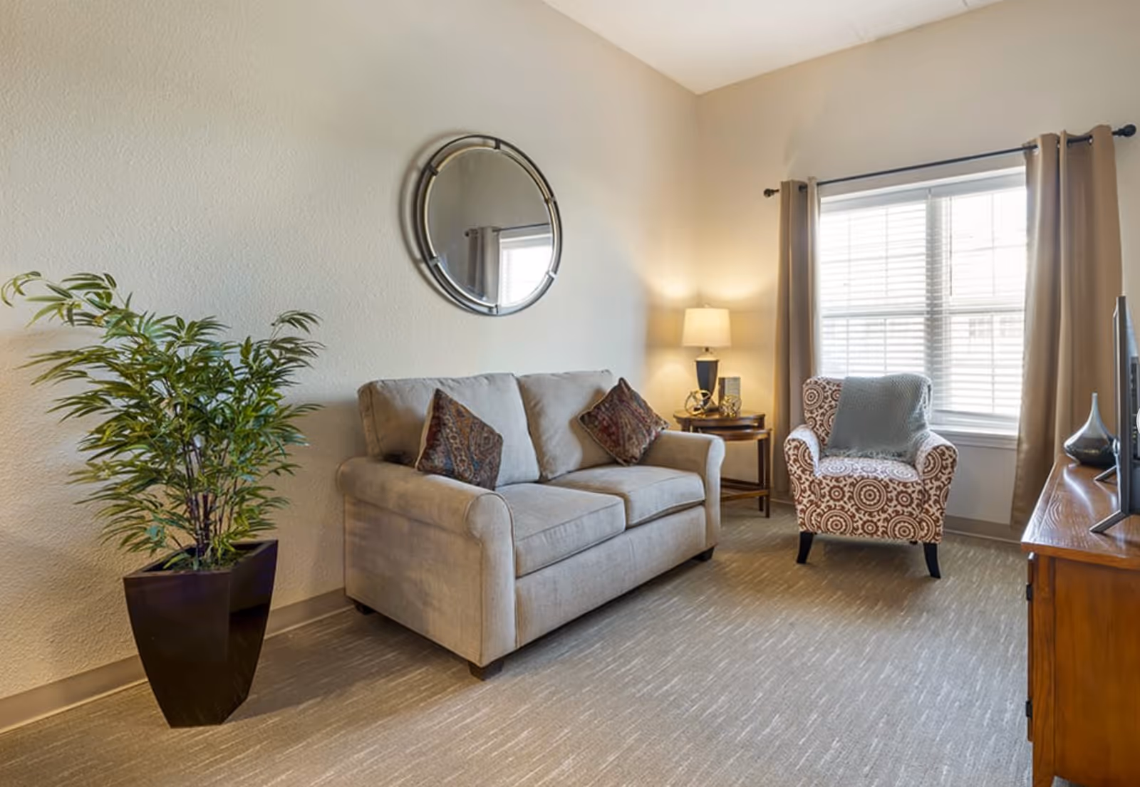 A cozy living room with a beige sofa adorned with two patterned pillows, a round mirror on the wall above the sofa, a potted plant to the left, a patterned armchair with a throw blanket near a window with beige curtains, a wooden side table with a lamp, and a wooden TV stand with a television.
