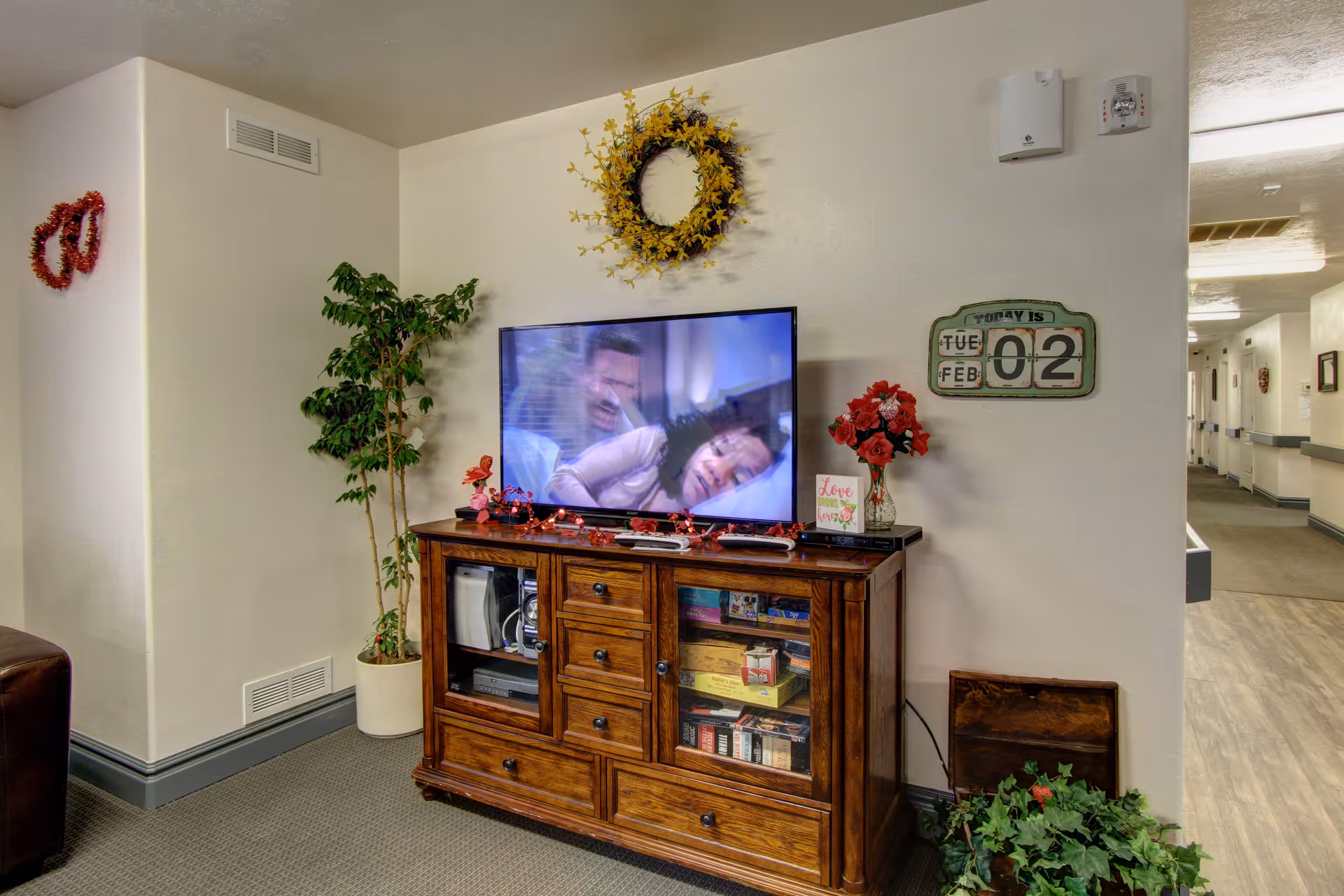 A TV on a wooden console in a decorated common area with potted plants, wall wreath, and a hallway visible to the right.