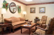 A cozy living room area with a striped sofa, two patterned armchairs, a wooden coffee table with decorative items, two side tables with lamps, a round mirror on the wall, and framed artwork. The room is warmly decorated with floral arrangements and has a beige and brown color scheme.