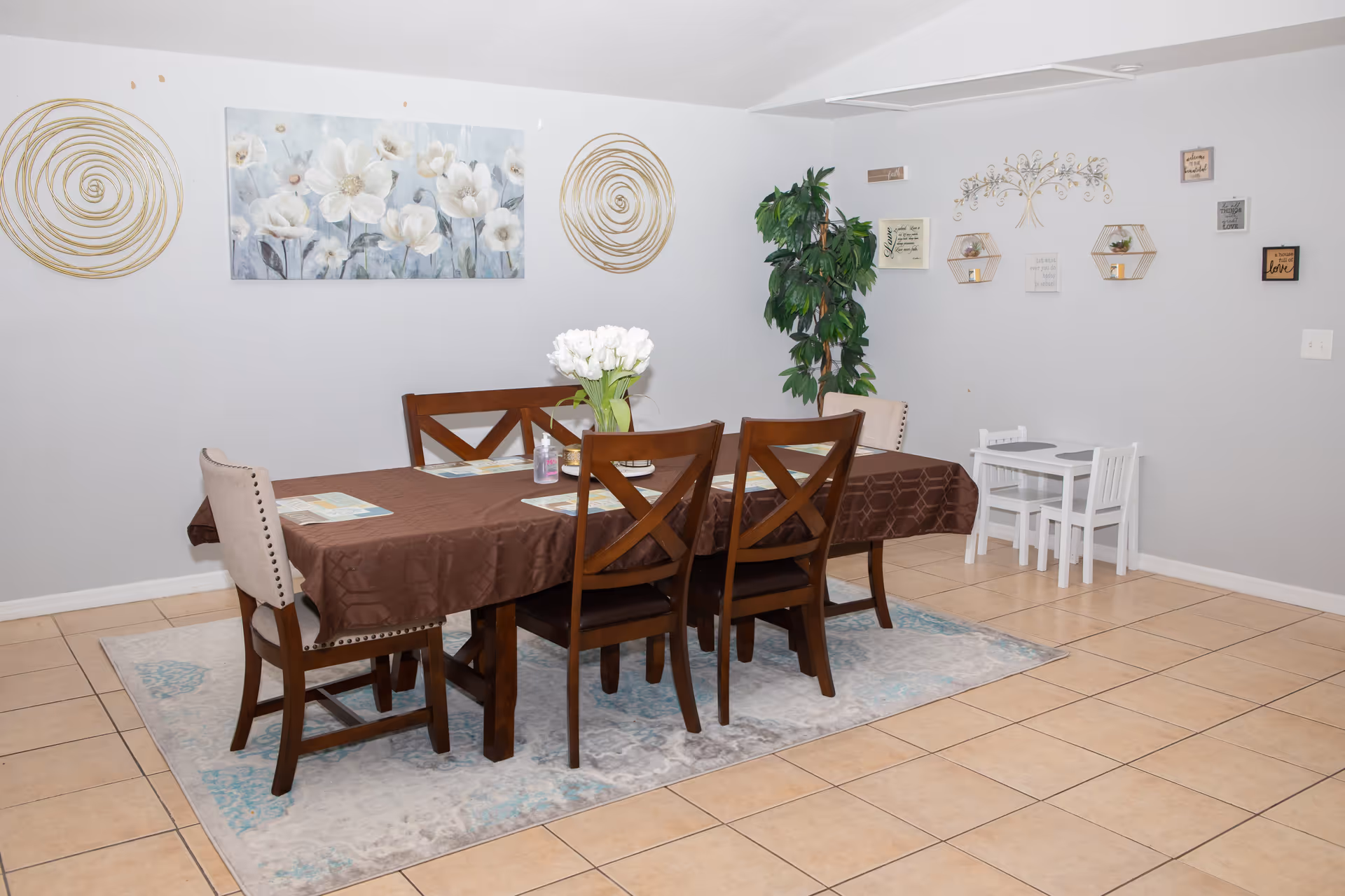 A dining room with a rectangular wooden table covered with a brown tablecloth and surrounded by six chairs, two upholstered and four wooden. The table has a vase with white flowers and placemats. The floor is tiled with a light-colored rug under the table. The walls are light gray and decorated with floral artwork, circular gold wall art, a green potted plant, and various framed quotes and decorative shelves. There is also a small white children's table with two chairs in the corner.