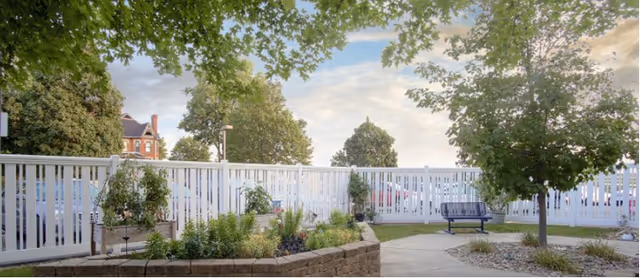 Outdoor garden area with a white picket fence, various plants and shrubs, a tree, a blue bench, and a paved walkway under a partly cloudy sky.