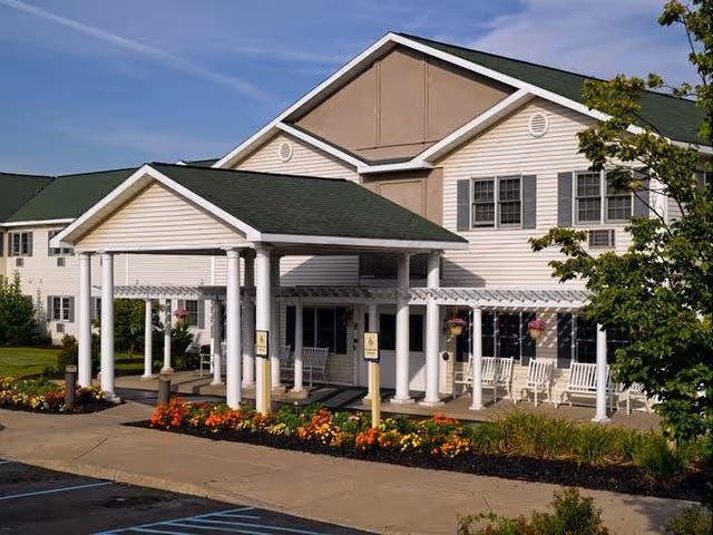 Exterior view of a senior living facility building with a covered entrance supported by white columns. The building has beige siding with white trim and a green roof. There are flower beds with colorful flowers along the sidewalk and several white rocking chairs on the porch. A tree is visible on the right side of the image under a blue sky.