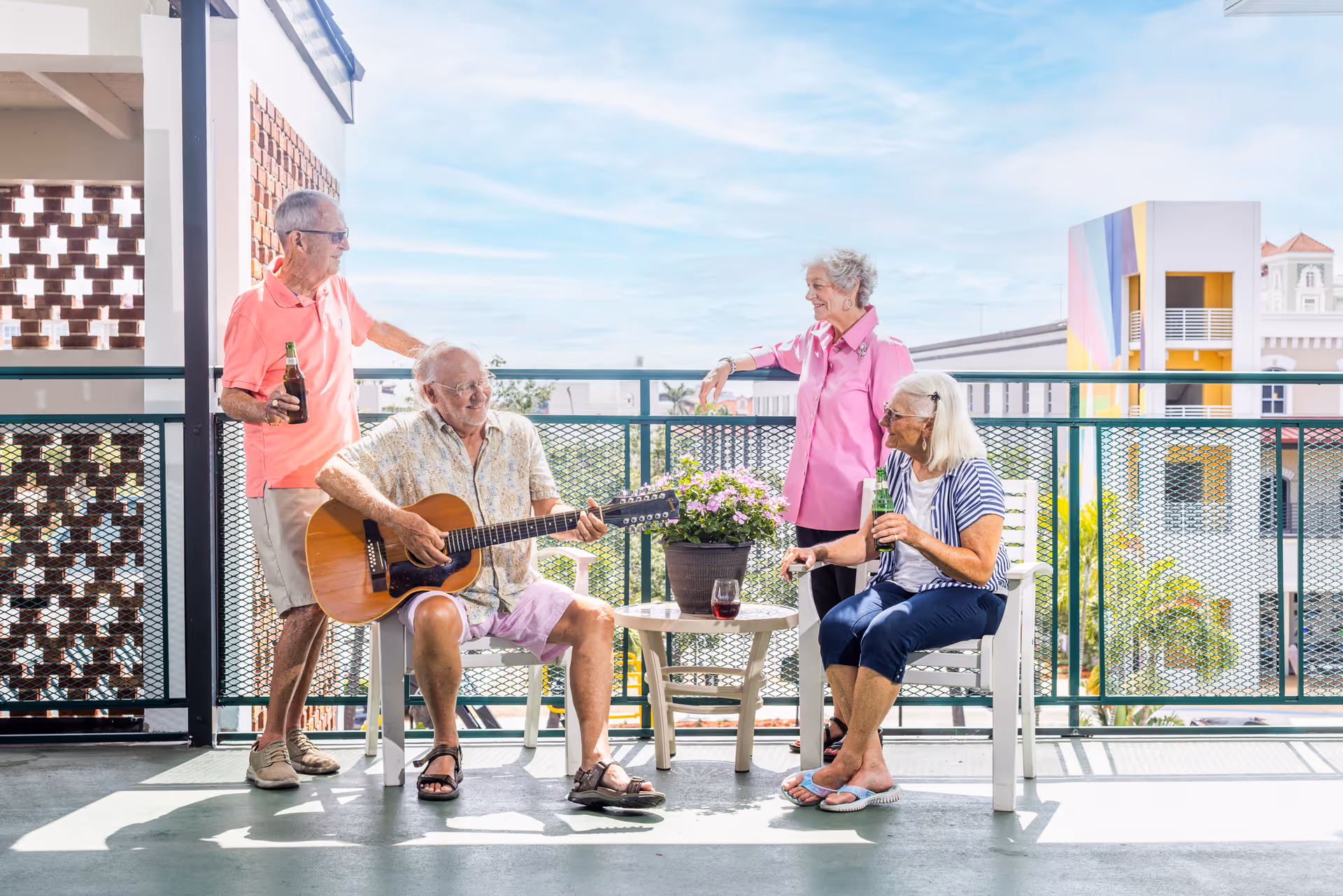 Four elderly people enjoying a sunny day on a balcony. One man is sitting and playing an acoustic guitar, while the other three, two women and one man, are standing or sitting nearby holding drinks and smiling. There is a small round table with a potted plant and a glass of red wine on it. The background shows a clear sky and colorful buildings.