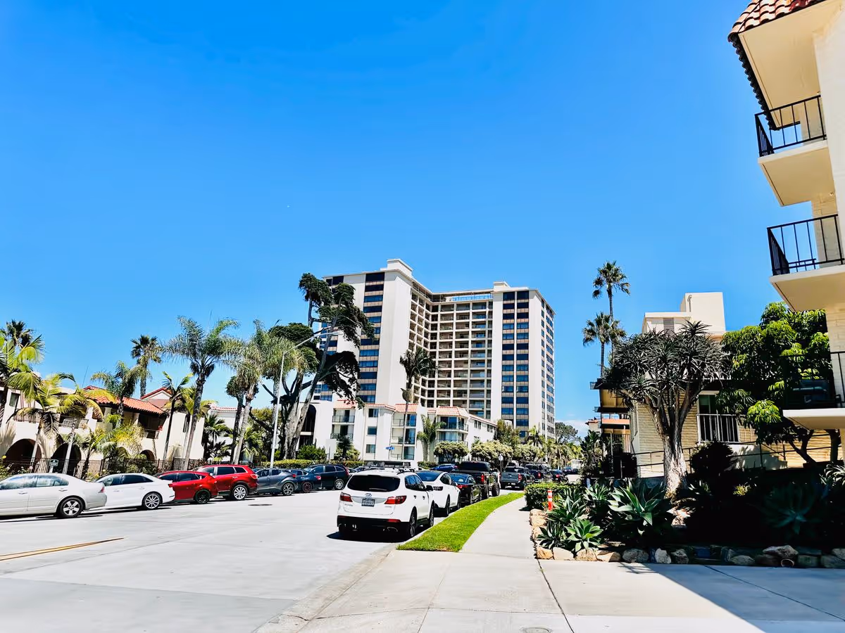 Street view of a multi-story retirement community building with palm trees, parked cars, and a clear blue sky.