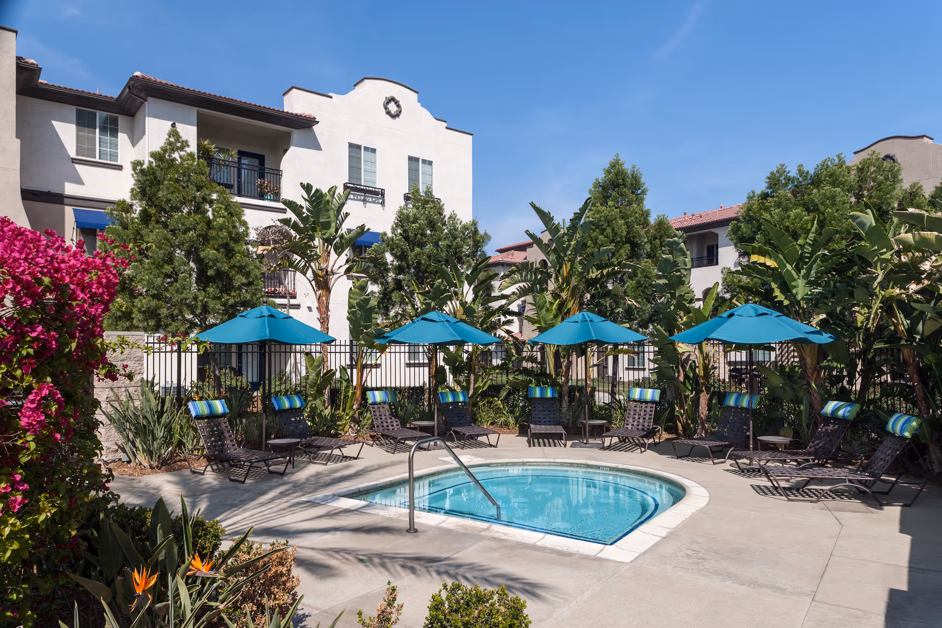 Outdoor pool area at Overture Riverwalk featuring a small round pool surrounded by lounge chairs with blue umbrellas. The area is landscaped with tropical plants and trees, with a multi-story residential building in the background under a clear blue sky.