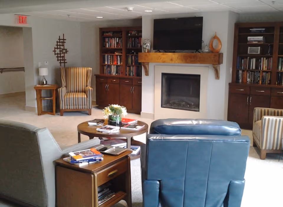 A cozy living room area with a fireplace, a wall-mounted TV above the fireplace, two wooden bookshelves filled with books on either side, a round wooden coffee table with books and a flower vase, and several armchairs including a blue leather chair, a striped fabric chair, and a gray chair. There is also a small wooden side table with books and a lamp, and an exit sign visible in the background.