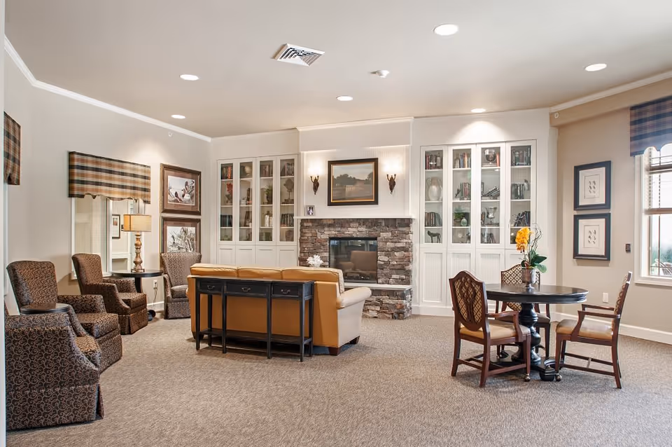 A cozy living room area in a senior living facility featuring a tan leather sofa facing a stone fireplace with a framed painting above it. On either side of the fireplace are white built-in cabinets with glass doors displaying books and decorative items. To the left, there are three patterned armchairs and a side table with a lamp. To the right, a round wooden table with four chairs is placed near a window with plaid valances. The room is well-lit with recessed ceiling lights and has neutral-colored walls and carpeted flooring.