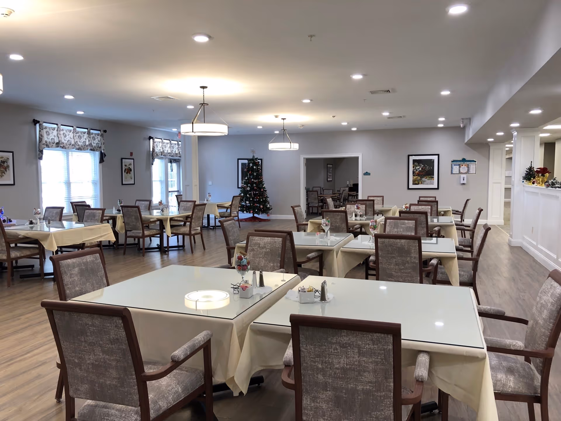 A spacious dining room with multiple tables covered with beige tablecloths and glass tops, each surrounded by cushioned chairs. The room has wooden flooring, soft lighting from ceiling fixtures, and windows with patterned valances. A decorated Christmas tree is visible in the background near a doorway leading to another room with more tables and chairs.