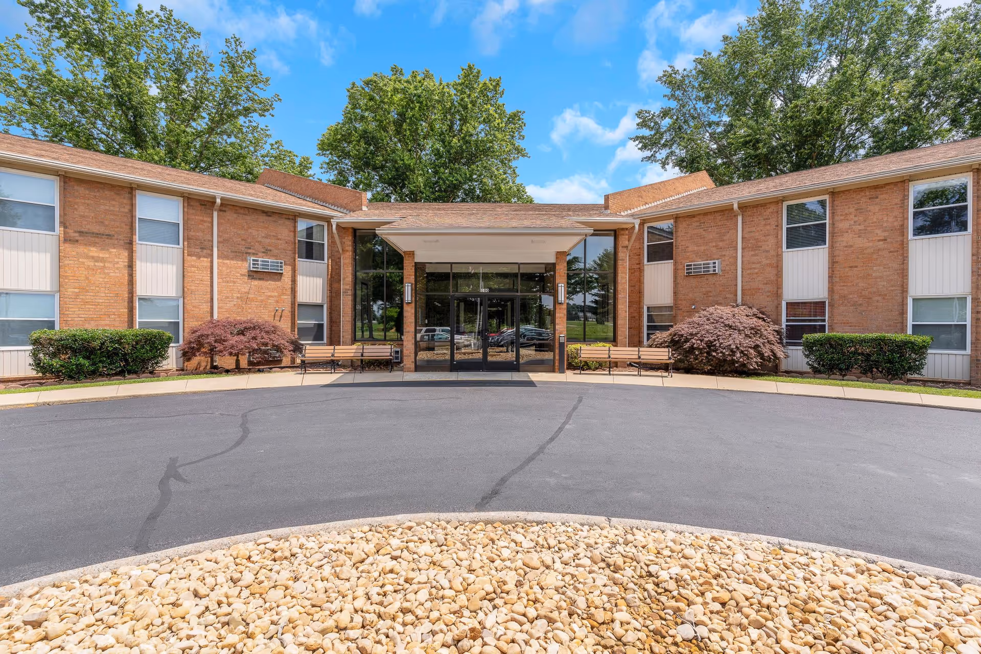 Front exterior view of a two-story brick building with large glass entrance doors, surrounded by bushes and trees under a blue sky with some clouds. There are benches on either side of the entrance and a circular driveway with a rock bed in the foreground.