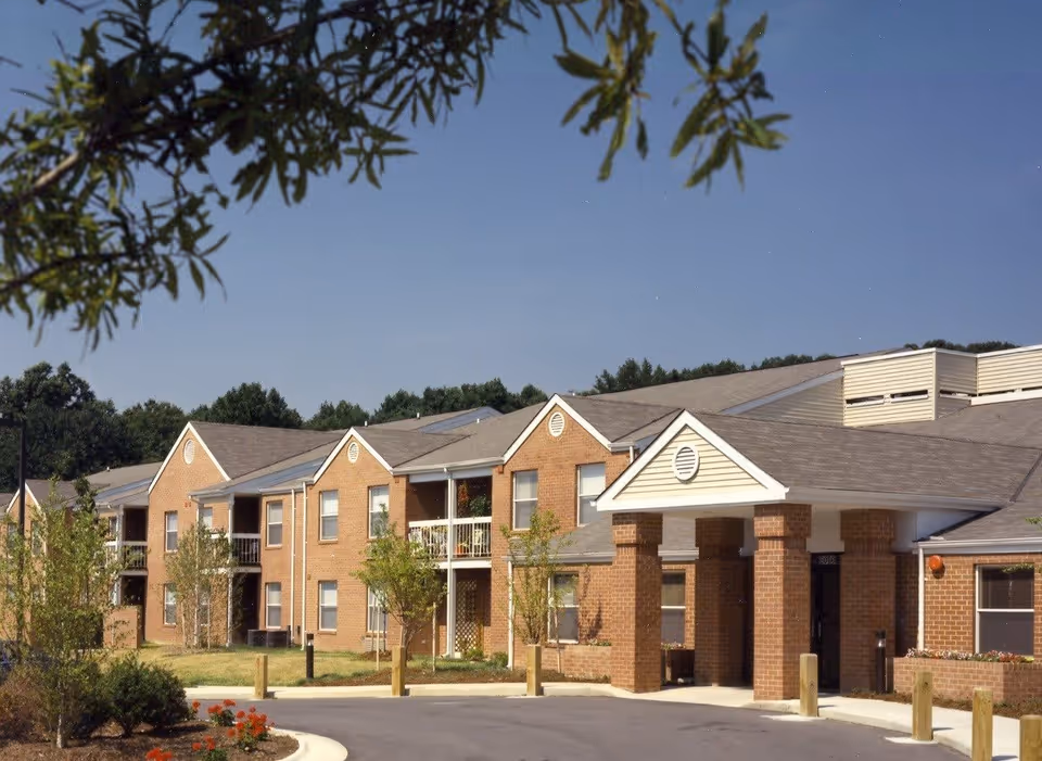 Exterior view of a two-story brick building with multiple windows and balconies, surrounded by small trees and landscaping under a clear blue sky. The building has a covered entrance supported by brick columns.