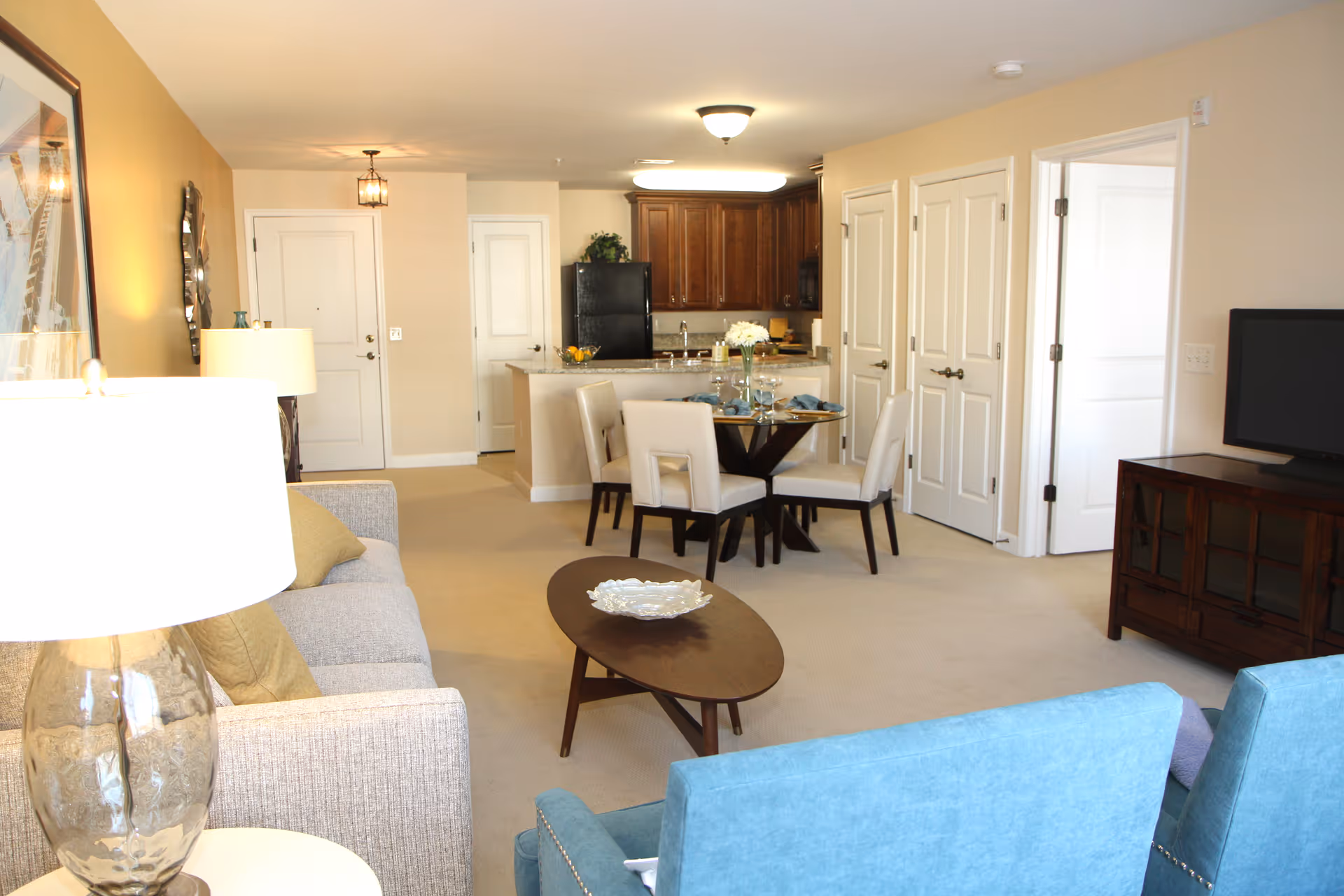 A bright and cozy living room and dining area in a senior living facility. The living room features a beige sofa with cushions, a glass table lamp, a wooden coffee table with a decorative bowl, and two blue armchairs. The dining area has a round wooden table set with plates, glasses, and napkins, surrounded by four white upholstered chairs. In the background, there is a kitchen with dark wooden cabinets, a black refrigerator, and a countertop with a bowl of fruit. The walls are painted in warm beige tones, and there are multiple doors leading to other rooms.