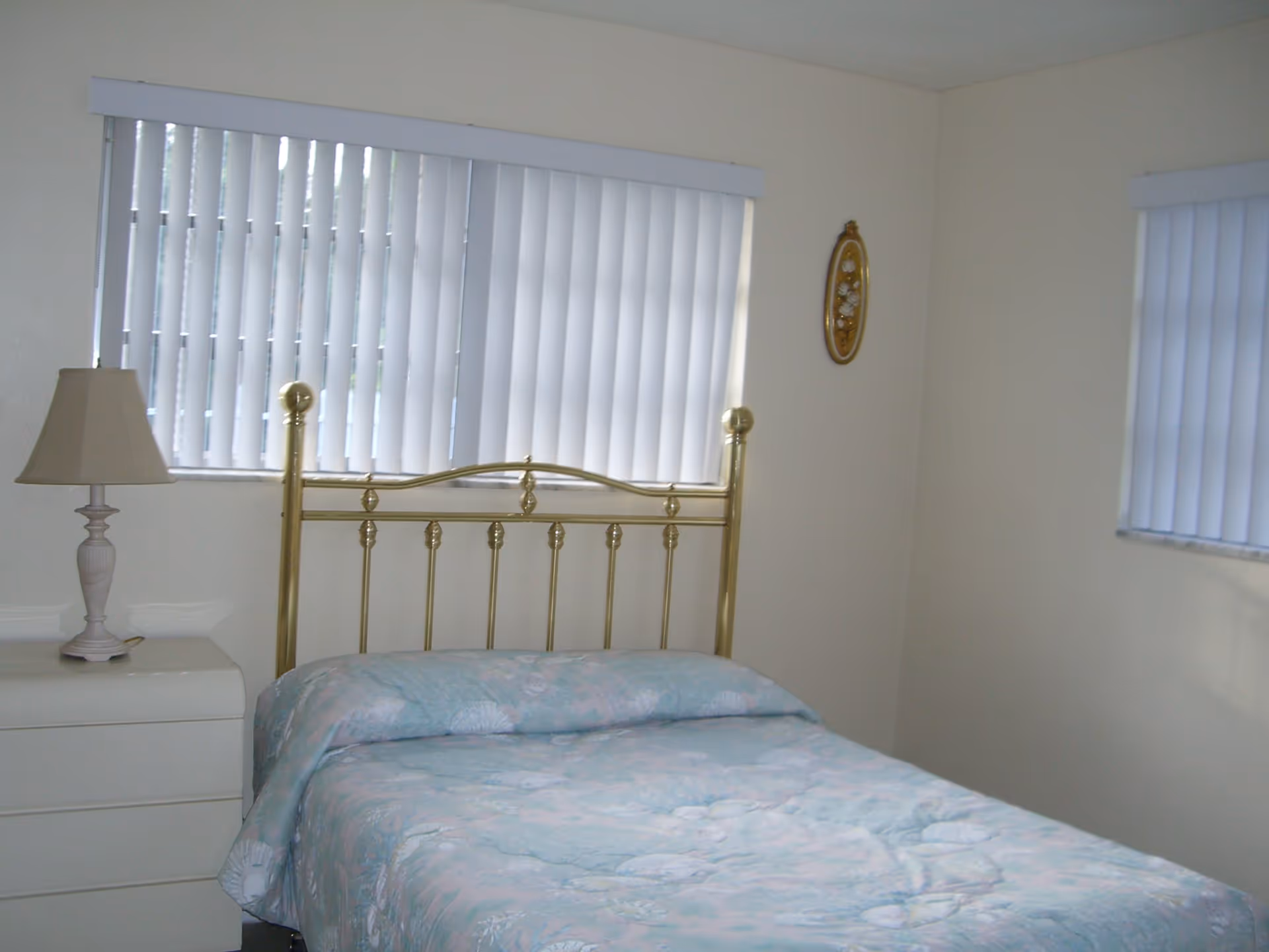 Small bedroom with a brass headboard bed, pastel bedspread, nightstand with a lamp, and vertical blinds on the windows.